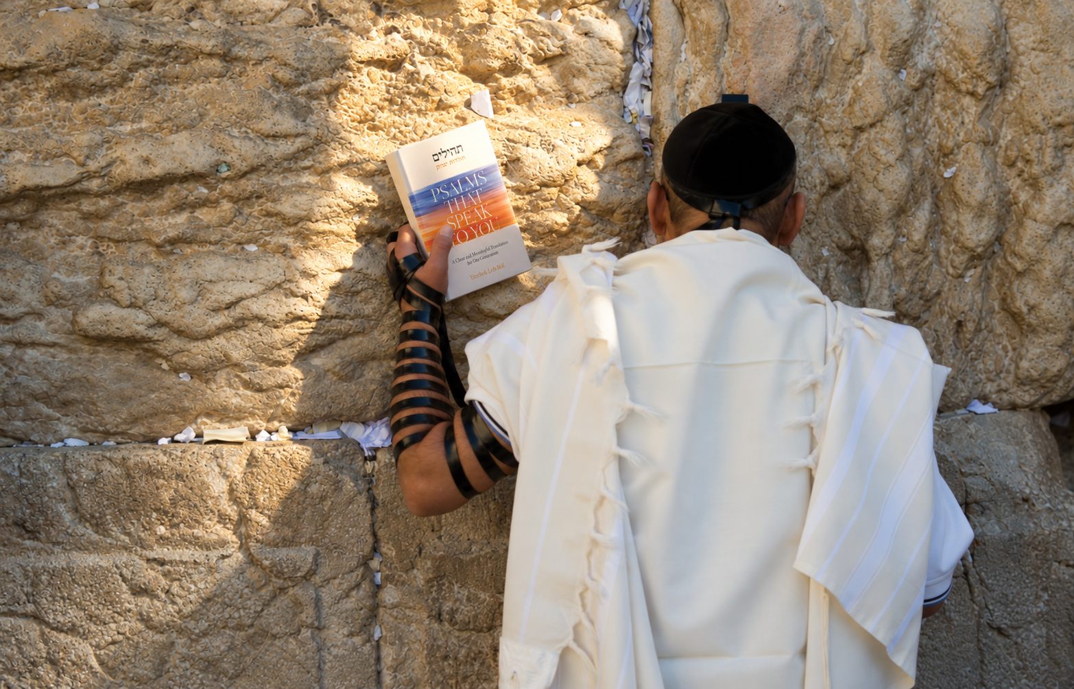 A man in tallis and tefillin holding Psalms That Speak to You against the stones of the Western Wall